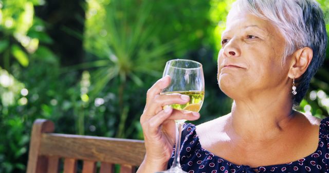A senior Caucasian woman enjoys a glass of wine outdoors, with copy space. Her relaxed demeanor and the natural setting suggest a moment of leisure or celebration.