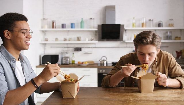 Friends enjoying takeout noodles at modern kitchen table using chopsticks, casual dining