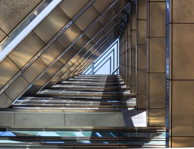 Featuring a striking geometric design, this photo showcases the modern architecture of what appears to be a building viewed from below, creating repeated triangular patterns with metallic surfaces. Ideal for use in design blogs, architectural websites, or marketing materials focused on contemporary urban development or abstract aesthetics.