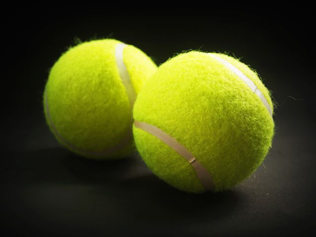 This close-up image of two bright yellow tennis balls on a dark background is perfect for use in sports-related content. It can be used for advertisements, promotional materials, or articles about tennis. The vivid contrast captures attention, making it ideal for any media requiring a focus on sports equipment.