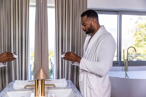 African american man in white robe examining skincare jar at marble sink and tub