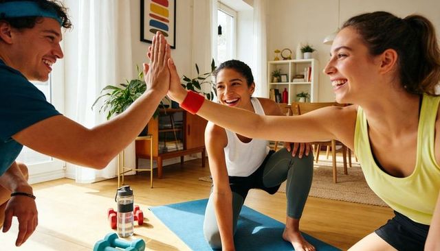 Friends high-fiving during home workout on yoga mat with dumbbells and hydration