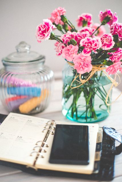 Floral-arranged desk scene with a planner, smartphone, and jar of pink carnations. Suitable for articles on organization, productivity, or lifestyle. Perfect for illustrating blog posts on workspace decor, daily planning, or managing schedules.