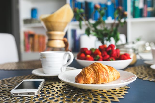 Appealing croissant on a white plate alongside a cup of coffee and a smartphone. In the background, strawberries in a bowl add a touch of color. Ideal for use in articles about breakfast ideas, modern kitchen decor, and casual dining settings. Great for blogs, advertisements, and social media posts related to lifestyle and food.