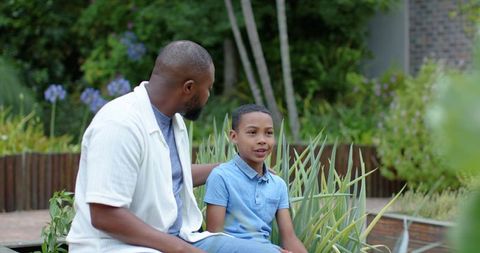 African american father and teenage son sitting and talking in garden with purple blooms