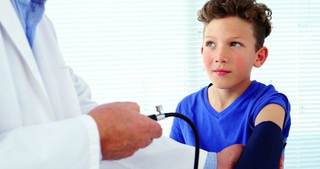 Doctor checking blood pressure of a young boy wearing a blue shirt in a clinical setting. The boy, seated and looking attentive, represents a routine medical exam. Ideal for use in healthcare advertisements, educational materials, pediatric care resources, and medical websites.