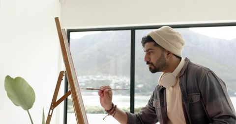 Indian artist painting at easel in bright mountain-view studio wearing headphones with lake view