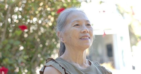 Senior asian woman smiling in sunny garden with red flowers and soft bokeh