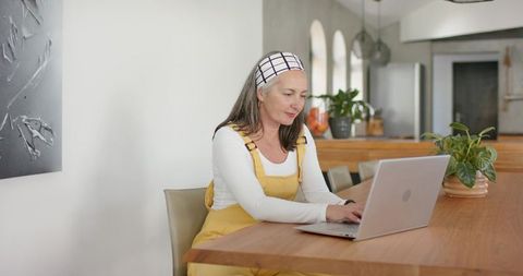 Mature woman working on laptop at wooden kitchen table wearing mustard overalls