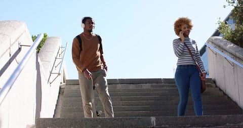 Two friends walking down urban stairs carrying skateboard and talking on phone in sunlight