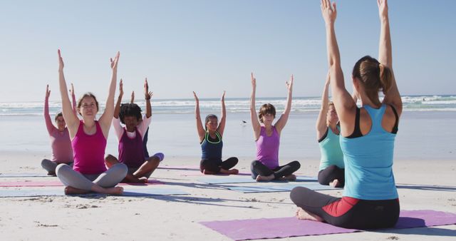 Group of people practicing yoga on beach, all sitting cross-legged with arms raised, clear blue sky and ocean in background. Great for promoting community fitness events, mindfulness retreats, health and wellness programs, or outdoor activities.