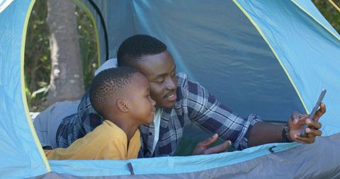 African american father and son bonding in tent, sharing smartphone while camping outdoors