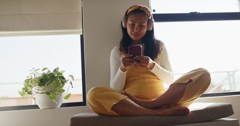 Mid-30s asian woman relaxing on sunlit window seat using smartphone and headphones