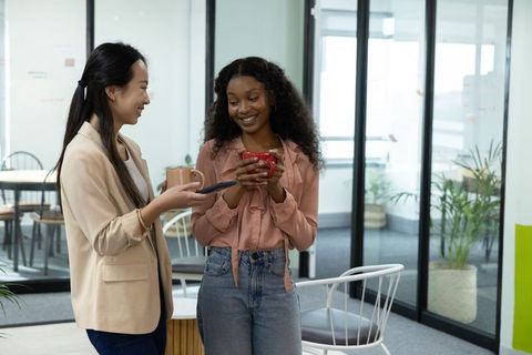 Diverse female coworkers chatting over coffee in modern glass-walled office lounge