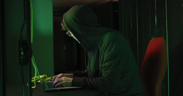 Asian male hacker in hoodie using laptop by computer servers. internet security, cyber crime, information technology, data processing and computer servers.