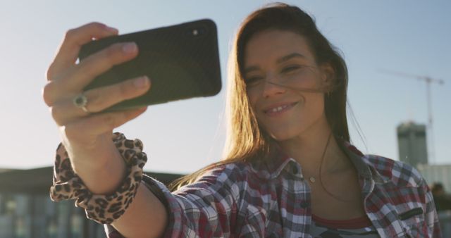 Young woman wearing a plaid shirt taking selfie with smartphone outdoors on a sunny day. Ideal for use in social media marketing, lifestyle blogs, fashion blog, technology ads or contemporary living themes.