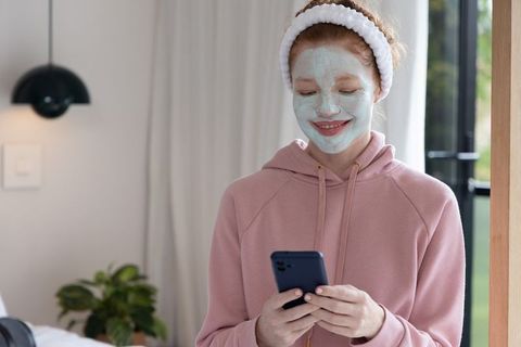 Teenager wearing facial mask and checking smartphone in cozy minimalist bedroom