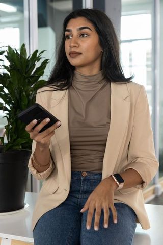 Indian businesswoman sitting on countertop holding smartphone and looking right in office