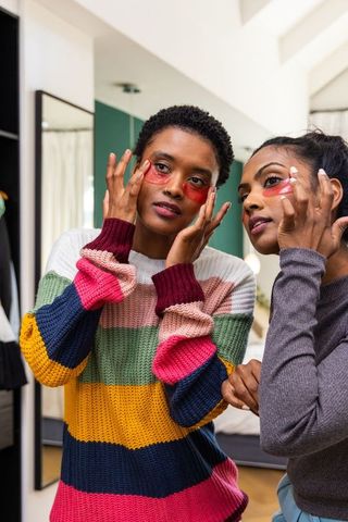 Multicultural friends applying under-eye gel patches in bedroom mirror, cozy sweaters
