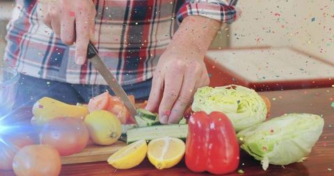 Male chef slicing cucumber on wooden board with fresh vegetables and confetti overlay