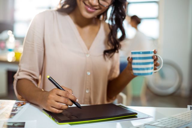 Businesswoman using graphic tablet while holding coffee mug in modern office. Ideal for illustrating concepts of multitasking, creative work, professional environment, and modern office settings. Suitable for use in articles, blogs, and marketing materials related to business, technology, and design.