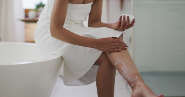 This visual showcases a woman in a white towel, applying lotion to her legs after taking a shower in a modern bathroom. The calm and clean aesthetic promotes concepts of self-care and skincare routine, making it ideal for use in advertisements related to personal care products like lotions, body creams, and self-care treatments. It can also be used in wellness blogs and articles focusing on daily routines and self-care tips.