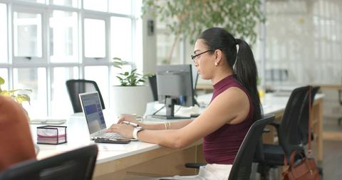Indian woman working on laptop at open-plan office desk, modern bright coworking workspace