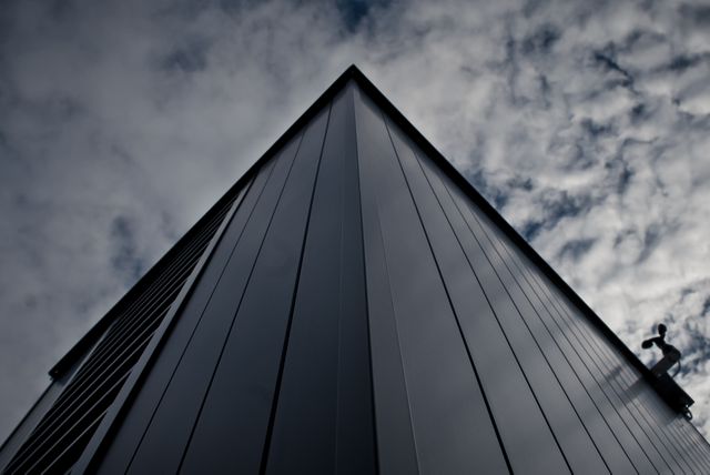 This stock photo captures a dramatic upward view of a modern building against a cloudy sky. The steel structure and sleek exterior design make this image ideal for architectural portfolios, construction marketing materials, and urban development presentations. The strong geometric lines and moody atmosphere highlight innovative architecture and can be used to emphasize concepts of progression, modernity, and cutting-edge design.