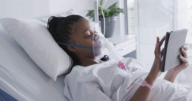 African American woman lying in hospital bed using a tablet device. She is wearing an oxygen mask. Scene shows a healthcare setting, highlighting use of technology in medical treatment and patient care. Suitable for content related to healthcare, digital health, patient care, recovery, and modern medicine.