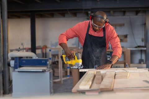 Skilled woodworker cutting plank with cordless jigsaw on workshop bench