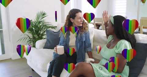 Two women chatting on sofa holding mugs with rainbow hearts overlay in sunlit living room