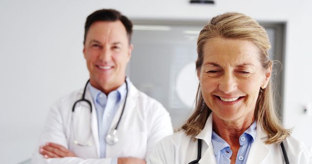 Two mature medical professionals, one male and one female, smiling in a hospital setting. Both are dressed in white lab coats with stethoscopes around their necks, suggesting confidence and teamwork. Ideal for illustrating healthcare, professionalism, medical services, or teamwork in medical environments.