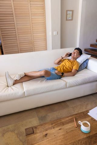 Asian man relaxing on white leather sofa talking on smartphone in minimalist living room