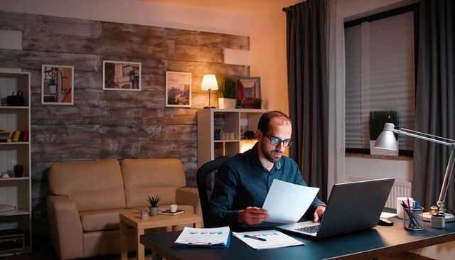 Man working late at home office, reviewing financial charts on laptop with reading glasses