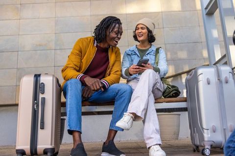 Laughing friends checking phone on transit bench with suitcases, urban travel lifestyle