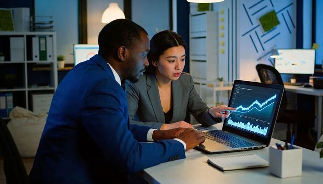 Colleagues analyzing rising financial charts on laptop in modern office at night
