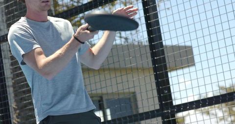 Man tossing flying disc on outdoor court with mesh fence and palm trees