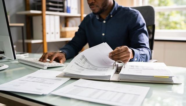 Business analyst reviewing financial reports, flipping binder and typing at desk