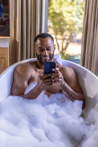 Mature african american man relaxing in bubble bath while texting by sunny window