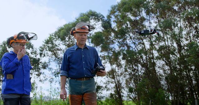 Forestry workers wearing safety gear operate a drone for forest management and surveillance. The workers use remote controls to navigate the drone in an open-space forest area, showcasing the integration of technology in environmental conservation efforts. Ideal for use in articles about modern forestry techniques, environmental monitoring, and the use of drones in various industrial applications.