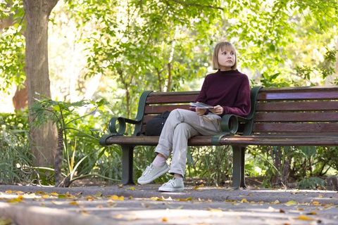 Young woman sitting on park bench reading booklet under autumn sunlight, quiet mood