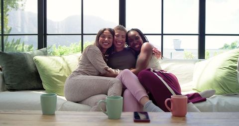 African american friends hugging on cozy sofa in sunlit living room with mugs and phone