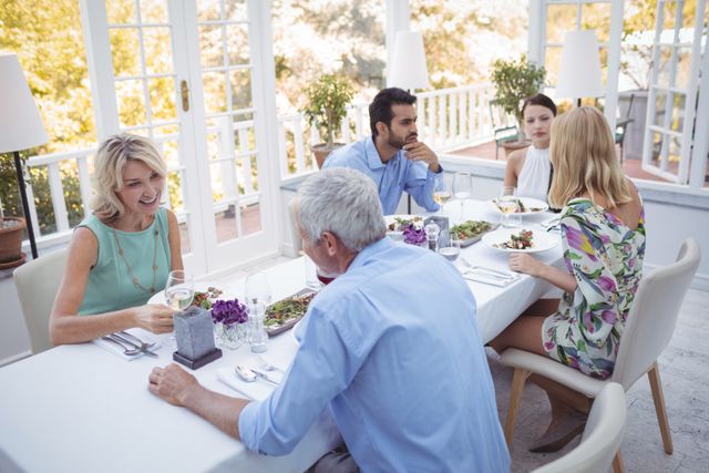 Group of friends enjoying a meal together in a bright and airy restaurant. They are engaged in conversation and appear relaxed and happy. This image is perfect for use in advertisements for restaurants, social gatherings, lifestyle blogs, and articles about dining out or social interactions.