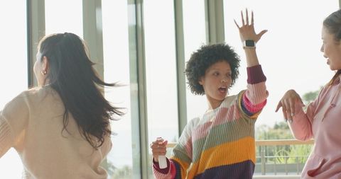 Young women dancing on balcony wearing colorful sweater and smartwatch, celebrating outdoors