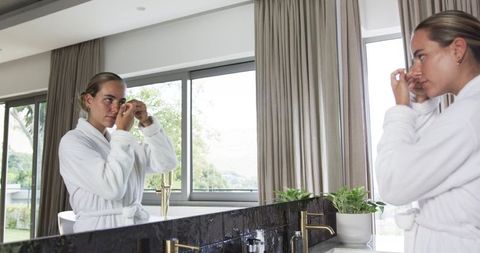 Woman in white bathrobe adjusting earring at luxurious marble bathroom vanity