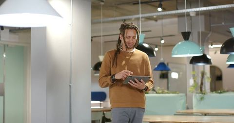 Young man with dreadlocks using tablet in modern coworking lounge under pendant lamps