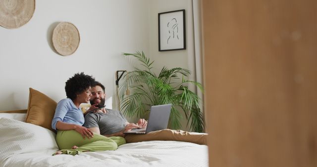 Image of happy diverse couple relaxing at home, lying on bed using laptop and smartphone, copy space. Happiness, communication, inclusivity, free time, togetherness and domestic life.