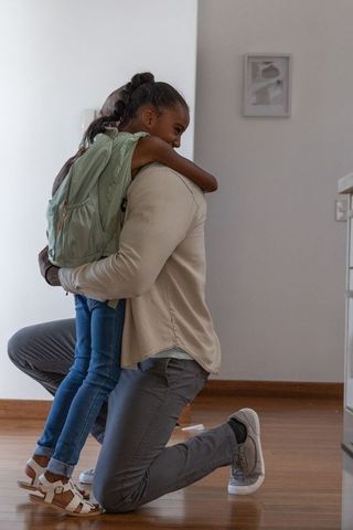 African american guardian hugging schoolgirl with light green backpack in home hallway
