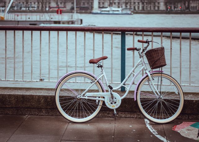 Vintage style white bicycle with brown leather seat and a front basket parked by a riverside pathway with a railing. The river and part of the city skyline are visible in the background, depicting urban life and transportation. Ideal for use in travel blogs, urban lifestyle content, transportation topics, and retro-themed projects.