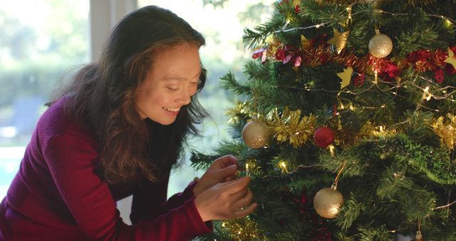 Woman decorating Christmas tree with various ornaments and lights, signifying festive holiday spirit. Ideal for use in articles and promotions about Christmas celebrations, home decor, or holiday-themed marketing materials.